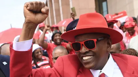 Mokgweetsi Masisi dressed in a red suit and wearing a red hat and sunglasses holds his fist up as he is surrounded by supporters.