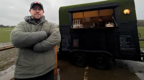 David Lamb, who is wearing a grey puffer jacket, stands with his arms crossed in front of a black converted horse trailer, which is now an independent coffee van. In the background area fields which form Herrington Country Park.