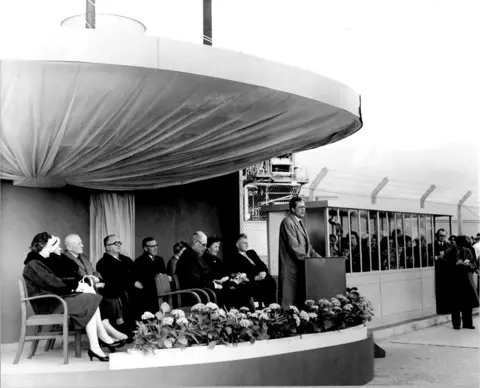 Courtesy Nuclear Restoration Services A black and white picture of the opening of the Chapelcross plant. A man is speaking at a platform with various dignitaries sitting behind him.
