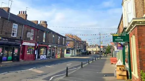 Bishopthorpe Road in York, filled with an array of different businesses on each side of the road, and there is bunting over the top of the buildings and on the road.