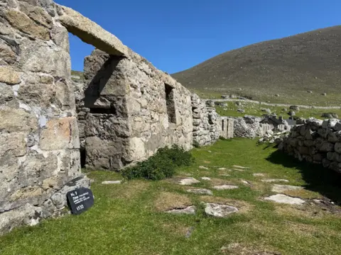 David May A derelict cottage on St Kilda on a sunny day - a small sign in front of the derelict building gives the details of the building