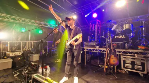 The Vanlife Festival A man who is wearing a black t-shirt and jeans is standing on a music stage. He is playing a guitar and singing into a microphone. Multi-coloured flashing lights surround him.