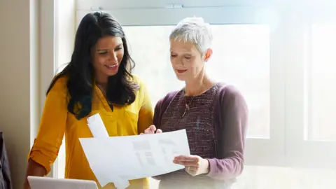 Two people talking stood reading a piece of paper. On the left a woman in a yellow blouse with dark brown hair and on the right a woman with a purple cardigan and short grey hair. 