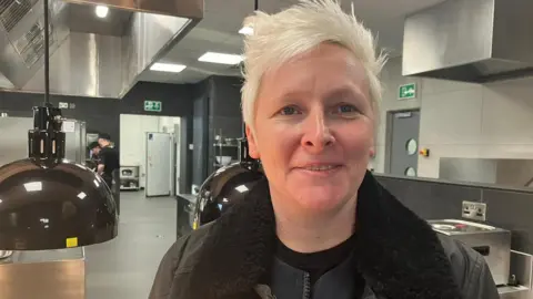 BBC/Gina Millson Lisa Goodwin-Allen in the new facility at Blackburn College. She stands in the kitchen by a hot plate and various other appliances. Lisa has short bleached blonde hair and is wearing a black jacket.