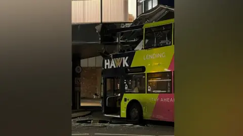 A lime green, black and pink double-decker bus crashed into a building overhang, with a crumpled top deck and debris lying in the road while it is still dark.