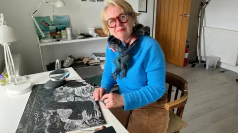 A woman wearing a blue jumper and a woollen scarf sits in an artist's studio. She is at a desk working on a black-and-white picture of puffins.