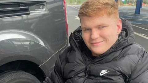 Ralph Pearce has short ginger hair and is wearing a black coat while sitting in a wheelchair in a car park. He is smiling.