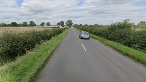 Google The B488 Leighton Road. A single grey car is travelling in the opposite direction and there are grass verges either side of the road