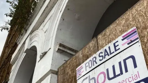 BBC For sale sign on a wooden board, positioned in between a white arch. 