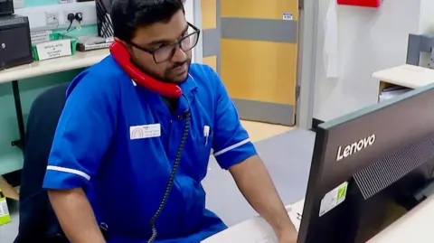 A male nurse is sitting at computer screen with a red phone pressed to his ear.  The man is about thirty years old and has been recruited to the NHS from Pakistan. He is wearing black rimmed glasses and has short dark hair and a short beard.