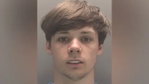 A mugshot of a young white man with hazel eyes and light brown hair. 