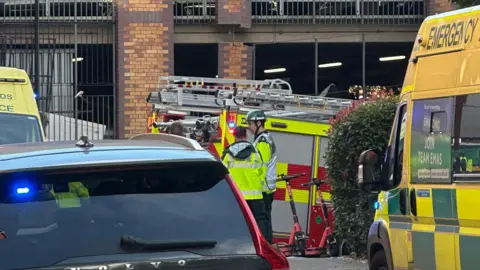 Jess Ramm Two ambulances, a first responder car and a fire engine in front of a brick-built multi-storey car park. Two emergency services personnel are visible in yellow hi-vis.