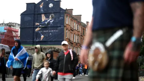 Reuters A large mural of Scott McTominay scoring an overhead kick against Denmark. The mural is painted on the side of a building wall. Football fans are walking past the wall.