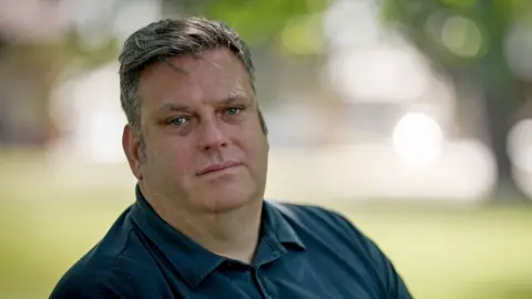 Steven is wearing a blue shirt and is sitting on a bench outside. He is in focus but the background, which resembles grass and trees, is fully out of focus. Steven is looking directly into the camera with a straight face. 