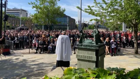 A reverend wearing a white cloak over a black robe is stood with his back to the camera and his head bowed. Hundreds of people are gathered in front of him and he is stood next to a memorial sculpture.