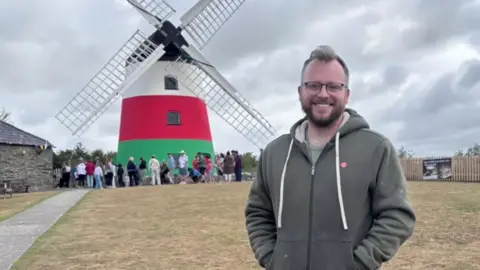 Richard Holt in front of a windmill painted red, white and green with people outside it. He has short blond hair and wears glasses.