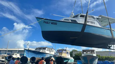 A blue-and-white fishing boat, the Pauly Wood, dangles from a crane against a blue sky.