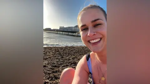 A woman takes a selfie by the sea with a pier in the background.