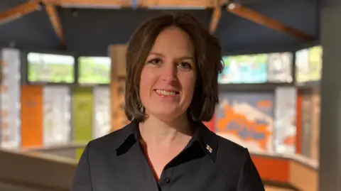 Josh Day/BBC Helen Glencross is smiling. She is standing in a museum and display boards can be seen in the background.
She has brown hair and is wearing a black blouse with a small pin broach on her lapel.