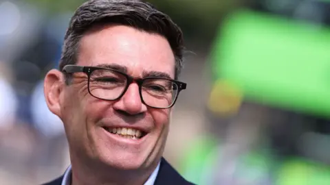 Andy Burnham is smiling and looking to the side. He has dark hair, brushed to one side and dark glasses.