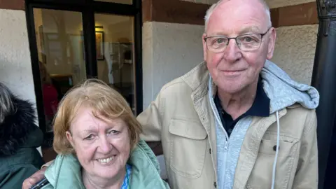 Cath and Peter Douthwaite, who are an older married couple. She is  wearing a green down jacket and blue blouse and has fair hair. He is wearing a khaki-coloured coat and a grey hoodie, is balding and wears glasses. 