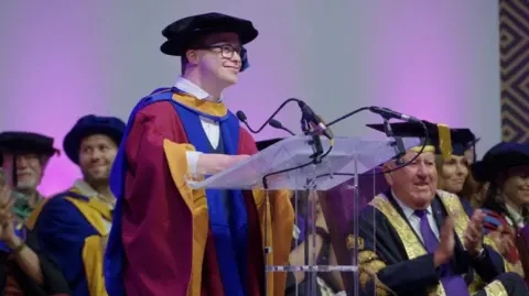 A picture of George Webster giving a speech at the graduation ceremony. He is wearing his red, gold and blue graduation robes and is standing at a lectern. There are people sat in their graduation robes behind him. 