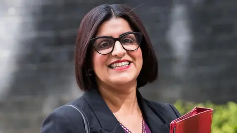 Getty Images A smiling Shabana Mahmood stood in front of a black brick wall and a green bush in front. She is wearing a black jacket, purple top, and is holding a red folder.