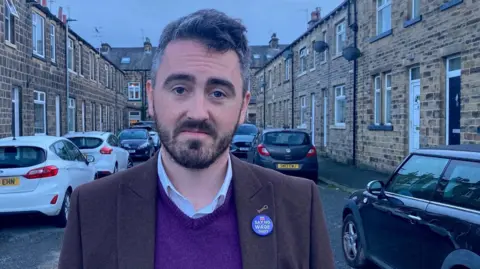 A man wearing a brown jacket and purple V-necked jumper standing in a street.