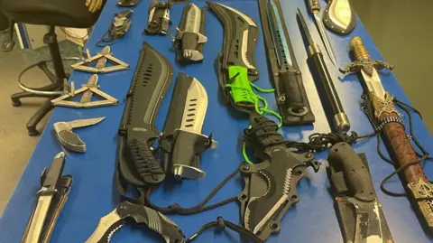 A blue table displaying a variety of bladed articles including machetes, large and small knives, lock knives and a sword.