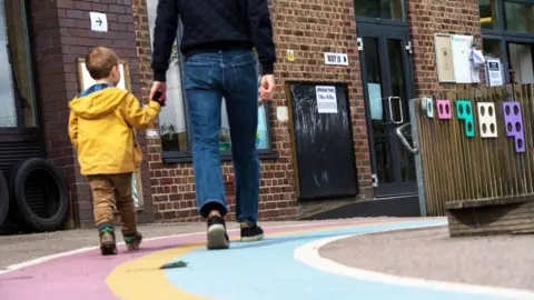 A small boy wearing a yellow raincoat holds his dad's hand as they walk into a polling station in a primary school.