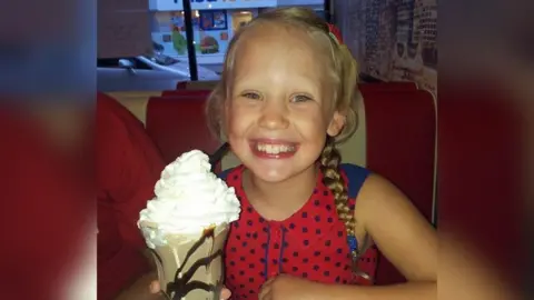 Family handout A young girl with a wide teethy smile sat in a red diner chair in a cafe holding a large ice-cream sundae. She has blonde hair in plaits and a red and blue polka-dot top on.