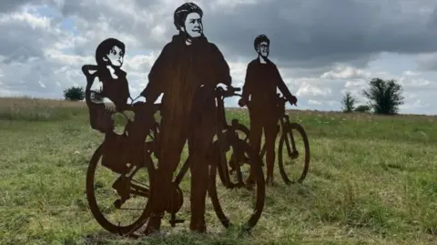 Steel statues of silhouettes on Salisbury Plain of a woman wearing a bicycle helmet holding a bicycle with a small child in a bike seat. A man holding a bicycle is in the background.