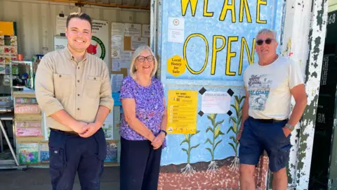 Sue Kinnear Three people smiling widely with a sign which says 'we are open'