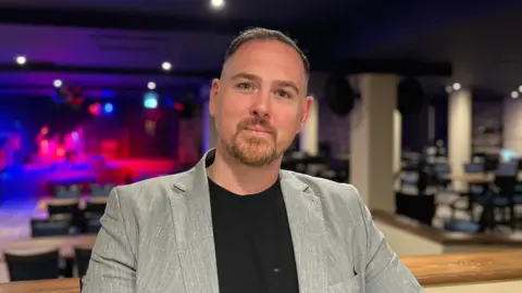 A man in a grey suit jacket and black tshirt pictured in a bar. A stage is visible in the background, with mood lighting. He has short brown hair and a goatee and is smiling. 