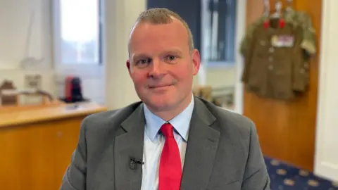 Kris Rotchell smiling at the camera, wearing a pale blue collared shirt underneath a grey blazer and red tie. He is sitting in an office space with a military uniform hanging on the wooden door behind him. 