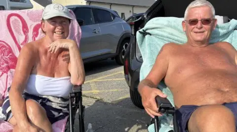Andy Gill/BBC Pauline Stain has a white sunhat and white tokp. Richard HOwared has white hair sunglasseds and blue shnorrts. Both arwe seated in deck chairs.