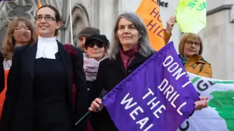 Getty Images Environmental campaigner Sarah Finch in 2019, having won her court case. She holds a purple flag with Don't Drill the Weald written in white on it, and is standing beside her female lawyer, who is wearing court robes.
Other campaigners stand behind them on the steps of the High Court.