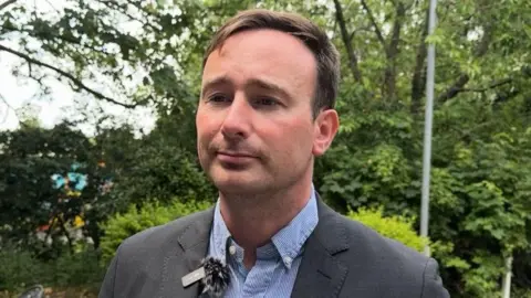 Tom Hayes MP being interviewed for BBC News. He is wearing a grey suit, no tie and a blue and white shirt. A hedge can be seen behind him.