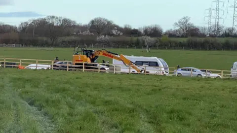 supplied A field with a wooden fence through the middle. On one side of it, there is a yellow digger and a caravan along with several cars.