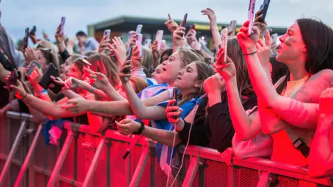 Dumfries and Galloway Council A crowd of young people at the barrier at an open-air gig. Many of them are filming a performance on their phones. There is a red tinge to the photo
