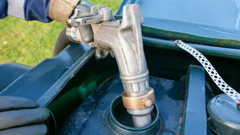 A silver pump is unloading a home heating oil delivery into a green plastic tank. You can see a person holding the pump. They are wearing a grey rubber glove with a blue and white sleeve.