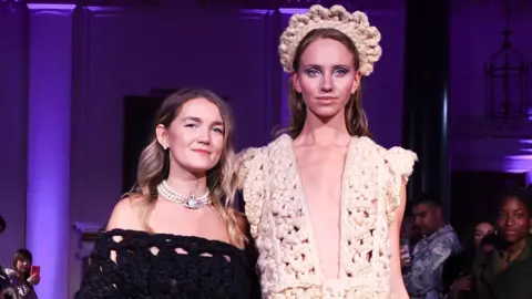 Getty Images Shonagh Murray (left) with a model wearing one of her designs. Shonagh has long blonde hair which has been curled and wears a black knitted off-the-shoulder dress and a pearl Vivienne Westwood necklace. The model on the right is considerably taller than Shonagh and wears a low-cut cream knitted dress with a matching crown-style headband. The stage behind them is lit purple. 