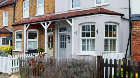 A row of terraced houses with brick and painted facades, white-framed windows, small front gardens, and low wooden fences along a residential street.