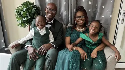 BBC A woman with glasses, a necklace, a long green dress, and a man with glasses and a green tuxedo, sit on a light grey sofa with their son, who is wearing a green waistcoat and suit, and daughter, who is wearing a green dress. All four of them are smiling. A plant is behind the man's right shoulder. Curtains behind them are grey and have white stars on them.