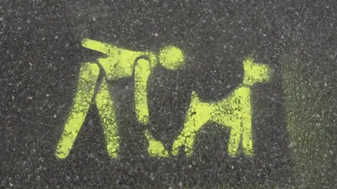 A yellow sign painted on a pavement shows a person cleaning up after their dog