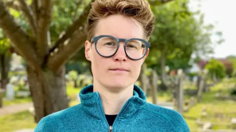Head and shoulders photo of Councillor Anna Railton with graves and trees in Wolvercote Cemetery, Oxford in the background. She is urging people to make plans and put money aside, to avoid "strangers" deciding what happens to them when they die.