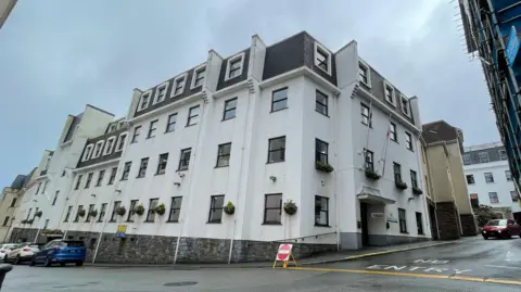 A view of Edward T Wheadon House. The five storey white building from a corner view. Cars are parked outside. 