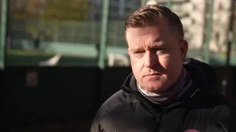 Eamon Gately in a black coat. He stands in front of a football pitch. 