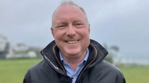 Man smiling at the camera wearing a wet weather jacket and a blue and white gingham shirt 