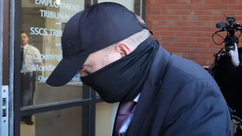 PA Media Robert Bush is wearing a navy coat, purple tie, black face covering and cap. He is entering a Hull Crown Court through an automatic glass door while keeping his head lowered. Behind him, several photographers and camera operators stand outside in front of a red brick wall, pointing their cameras toward him.